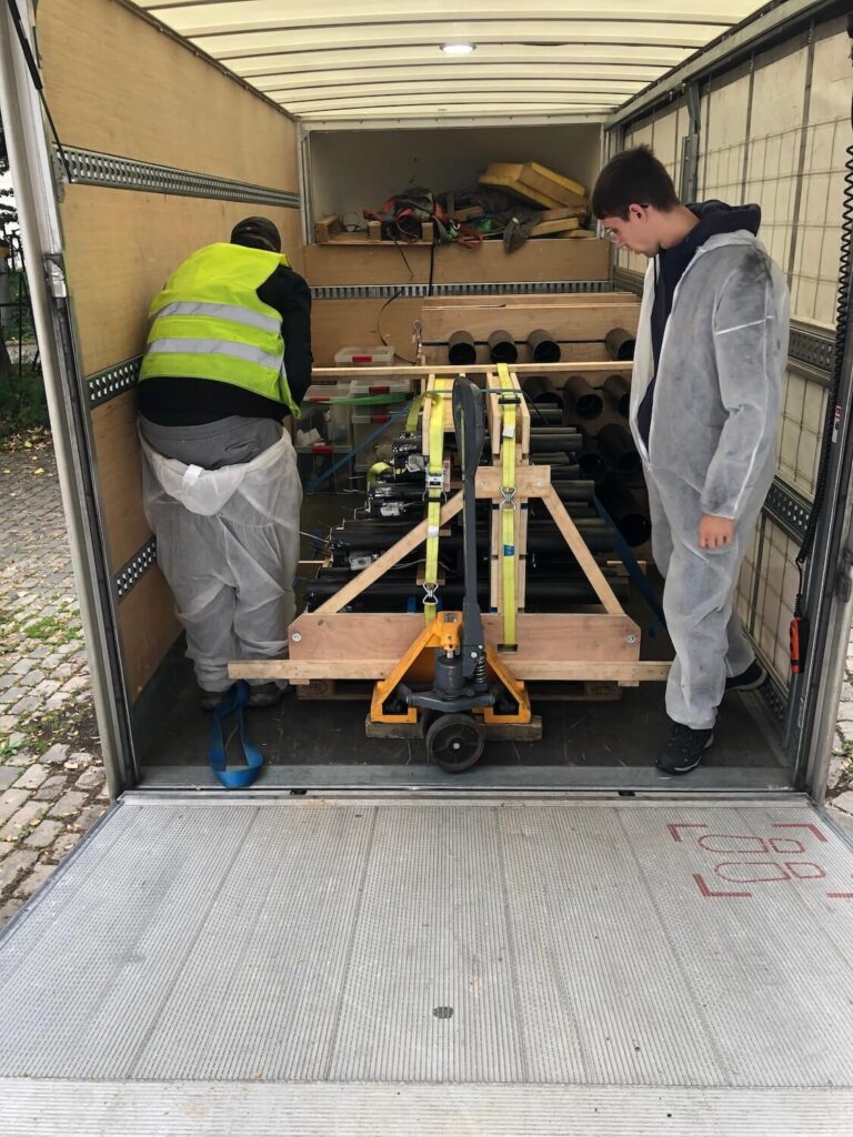 Photo montrant deux personnes en combinaison. blanche qui positionnent une palette contenant des cylindres noirs enchassés dans des structures en bois,-à l’arrière d’un camion